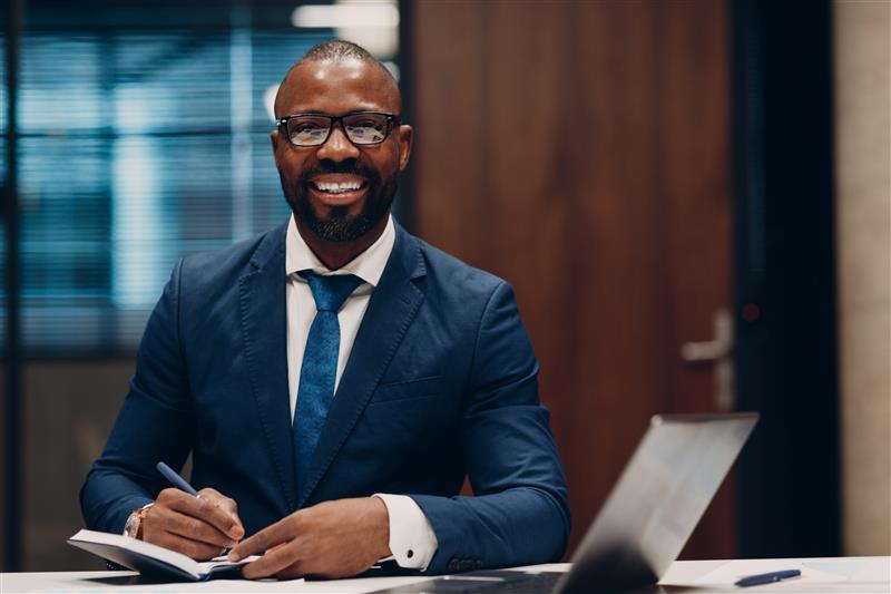 portrait smiling african american businessman blue suit sit table meeting office with notebook with pen laptop